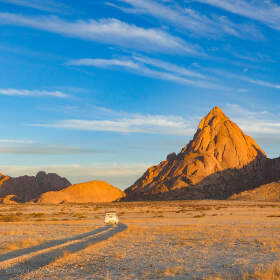 camping car driving on track through rocky plain with dry grass & red hills at sunset