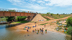 people bathing and washing clothes in river next to bridge